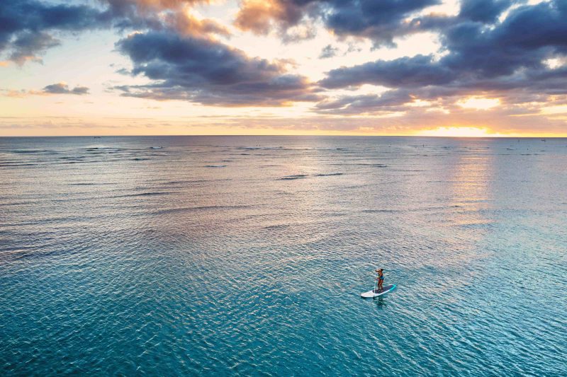 Aerial view of a paddle boarder paddling in the ocean during sunset.