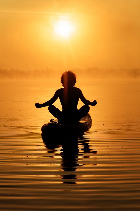 Silhouette of relaxed man sitting in lotus pose on paddle board with beautiful sunrise behind. 