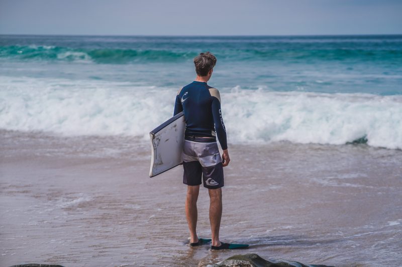 Middle aged male standing on the shore break holding his boogie board staring towards the surf.