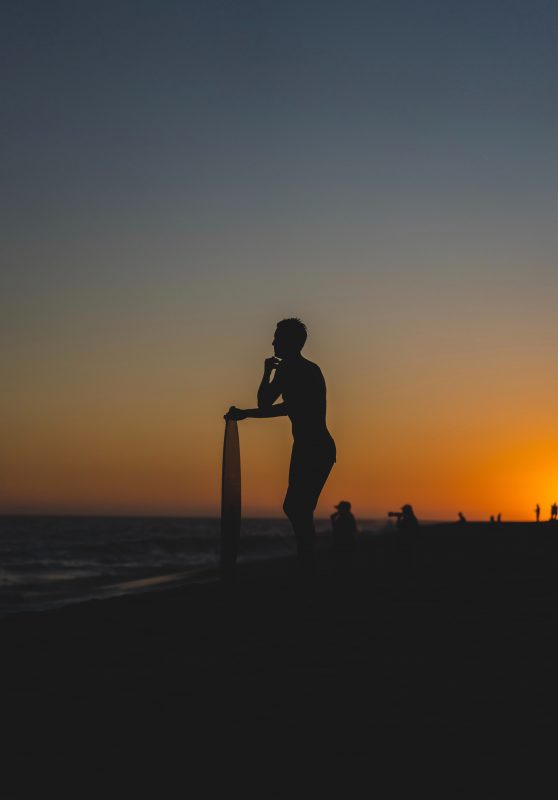 Skim Boarder resting on his board while looking out on the water during the sunset.
